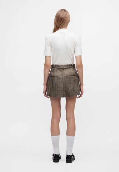 Femme debout de dos, portant un haut blanc à manches courtes, une mini-jupe marron rayée, des chaussettes blanches et des chaussures noires.