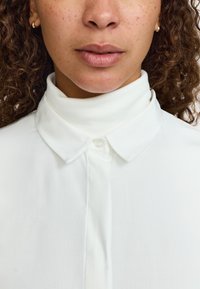 Camicia bianca con collo alto e tessuto a coste, dotata di chiusura con bottoni. Tessuto liscio con una leggera lucentezza.