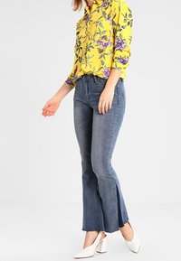 Camicia gialla a fiori con una vestibilità rilassata, abbinata a jeans svasati blu con spacchi laterali; le scarpe bianche con tacco a blocco completano l'outfit.