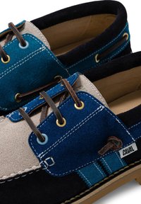 Gros plan sur des chaussures bateau en daim bleu, beige et noir avec des lacets en cuir marron et des détails de couture blanche.