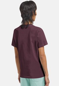 Persoon met halflang donker haar draagt een effen donkerpaarse t-shirt en lichtblauwe shorts, met de rug naar de camera gekeerd tegen een witte achtergrond.