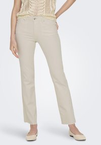 Donna che indossa pantaloni beige a gamba dritta con orlo grezzo, top in crochet crema e ballerine beige su uno sfondo chiaro e uniforme.