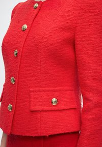Κόκκινο υφασμάτινο σακάκι με έξι χρυσά διακοσμητικά κουμπιά στο μπροστινό μέρος και ένα στη αριστερή τσέπη με καπάκι, φορεμένο με ταιριαστό κόκκινο παντελόνι.