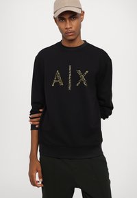 Sudadera negra con cuello redondo y mangas largas, que presenta letras doradas con tachuelas "AIX" en el frente. Textura de tela suave.