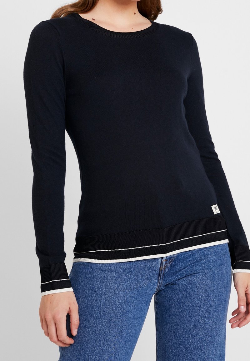 Femme portant un pull noir ajusté à manches longues avec des bordures blanches aux poignets et à l'ourlet, associé à un jean bleu, sur un fond uni.