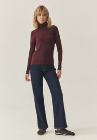 Femeie în picioare, îmbrăcată cu un pulover burgund cu guler înalt, blugi drepți de culoare albastru închis și adidași casual maro, pe un fundal uniform.