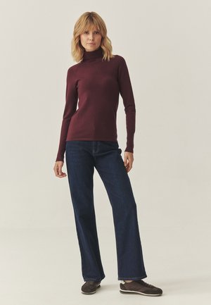 Mujer de pie con un suéter de cuello alto burdeos, jeans rectos de color azul oscuro y zapatillas casuales marrones frente a un fondo sencillo.