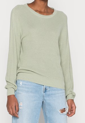 Personne portant un pull tricoté vert clair à manches longues et un jean bleu clair déchiré.