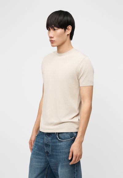 Νεαρός άνδρας με εφαρμοστό μπεζ πουκάμισο κοντομάνικο και μπλε τζιν, στέκεται πλάγια μπροστά σε απλό φωτεινό φόντο.