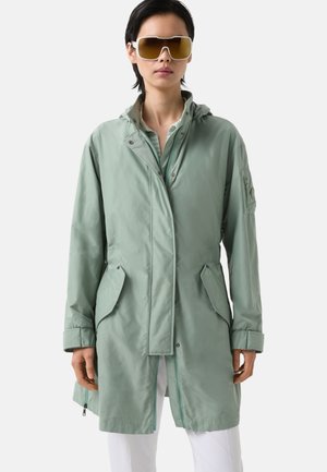 Veste imperméable vert clair avec capuche, dotée de deux poches avant, d'une fermeture éclair et d'une coupe décontractée. Des lunettes de soleil surdimensionnées complètent le look.