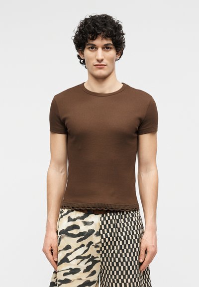 Νεαρός άνδρας με σγουρά μαλλιά, φορώντας καφέ στενή κοντομάνικη μπλούζα και σορτς με δύο μοτίβα σε μαύρο και άσπρο, σε απλό φόντο.