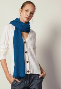 Écharpe bleue en matière douce drapée avec élégance ; associée à un cardigan blanc à côtes avec des boutons marron et un design en V.