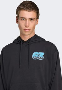 Haut à capuche noir avec une poche avant, arborant un graphique bleu "63" sur la poitrine. Le tissu semble doux avec une texture lisse et une capuche ajustable.