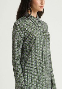 Camicia verde con bottoni, caratterizzata da un motivo geometrico viola e bianco. Maniche lunghe, colletto e tessuto morbido.