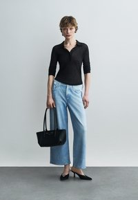 Femme portant un polo noir à manches longues, un jean large bleu clair, des talons pointus noirs, tenant un sac à main noir, debout devant un fond gris uni.