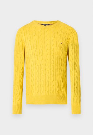 Sárga, kötött, hosszú ujjú pulóver, kerek nyakkivágással és kis Tommy Hilfiger logóval a mellkasán, fehér háttér előtt.