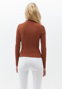 Langärmliges, enganliegendes Oberteil in Rostfarbe mit gerippter Textur, kombiniert mit weißen Jeans. Die Rückansicht betont die Silhouette und die Stoffdetails.