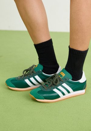 Stopy w zielono-białych butach sportowych Adidas z czarnymi skarpetkami, stojące na jasnozielonej podłodze.