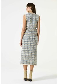 Vrouw in een mouwloze midi-jurk van blauw en beige tweed met franjes, gecombineerd met beige laarzen met een hak tot halverwege het onderbeen, staand met de rug naar de camera.
