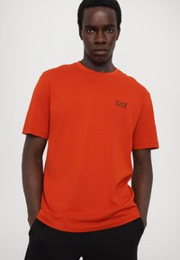 Tricou din bumbac portocalie cu gât rotund. Are un mic logo negru pe partea stângă a pieptului. Mâneci scurte și o croială relaxată.