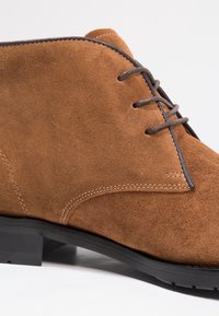 Bottine en daim marron avec un bout arrondi, des coutures décoratives et des lacets gris ; elle possède une semelle en caoutchouc noire pour la traction.