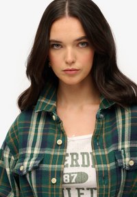 Chemise en flanelle à carreaux verts et marins avec fermetures à boutons, présentant deux poches poitrine et une texture douce, superposée sur un débardeur graphique blanc.