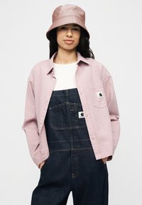 Γυναίκα που φοράει ανοιχτό ροζ καπέλο τύπου bucket, ροζ πουκάμισο με υφή και κουμπιά, λευκό μπλουζάκι και σκούρα μπλε τζιν σαλοπέτα με τα χέρια στις τσέπες.