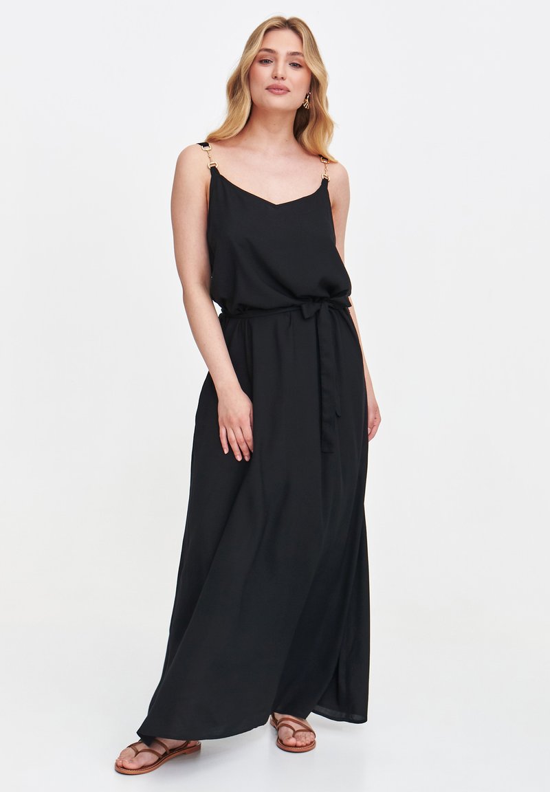 Kobieta stoi pewnie, ubrana w czarną sukienkę maxi na ramiączkach z wiązaniem w talii, w połączeniu z sandałami, delikatnie uśmiechając się do aparatu.