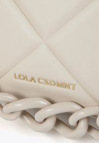 Beige textured leather bag with gold logo "LOLA CSDMNT" and a glossy cream chain detail at the bottom. Quilted design on the surface.