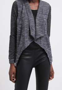 Stickad cardigan i svart och grått, öppen framsidan med draperad design, strukturerad tyg, långa ärmar, ihopparad med svarta åtsittande byxor.