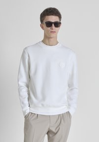Sudadera blanca con cuello redondo y mangas largas, con un detalle de flor texturizada en el lado izquierdo del pecho. Llevada con pantalones beiges.