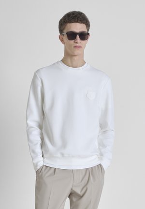 Sudadera blanca con cuello redondo y mangas largas, con un detalle de flor texturizada en el lado izquierdo del pecho. Llevada con pantalones beiges.