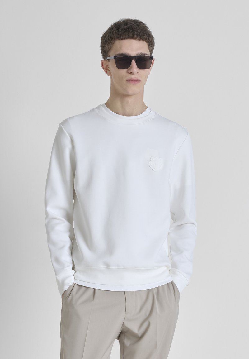 Sudadera blanca con cuello redondo y mangas largas, con un detalle de flor texturizada en el lado izquierdo del pecho. Llevada con pantalones beiges.