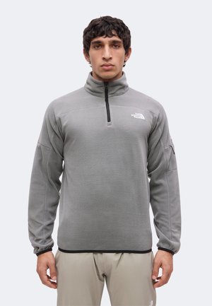 Homme portant une polaire gris clair avec fermeture éclair quart de zip et le logo The North Face sur la poitrine, ainsi qu'un pantalon beige, debout devant un fond clair uni.