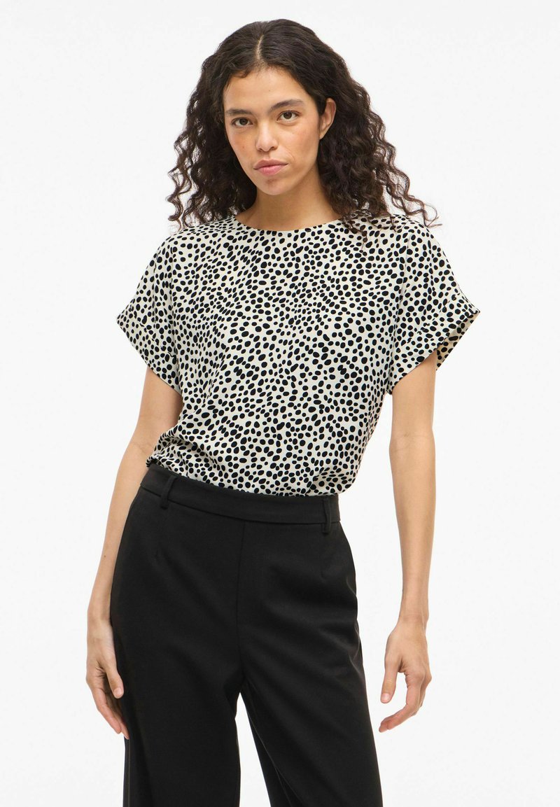 Femme aux cheveux bouclés foncés portant un chemisier blanc à manches courtes avec des pois noirs irréguliers et un pantalon noir taille haute sur fond blanc.