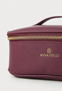 Neceser burdeos con superficie texturizada, cremallera dorada y logo de la marca. Forma rectangular compacta con un asa para llevar.