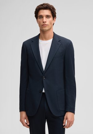 Homme aux cheveux bouclés portant une veste de costume bleu marine foncé sur un t-shirt blanc, sur un fond gris uni.