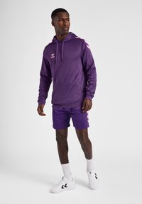 Felpa sportiva viola con dettagli e logo rosa, abbinata a pantaloni corti viola coordinati. Il modello indossa scarpe da ginnastica e calze bianche.