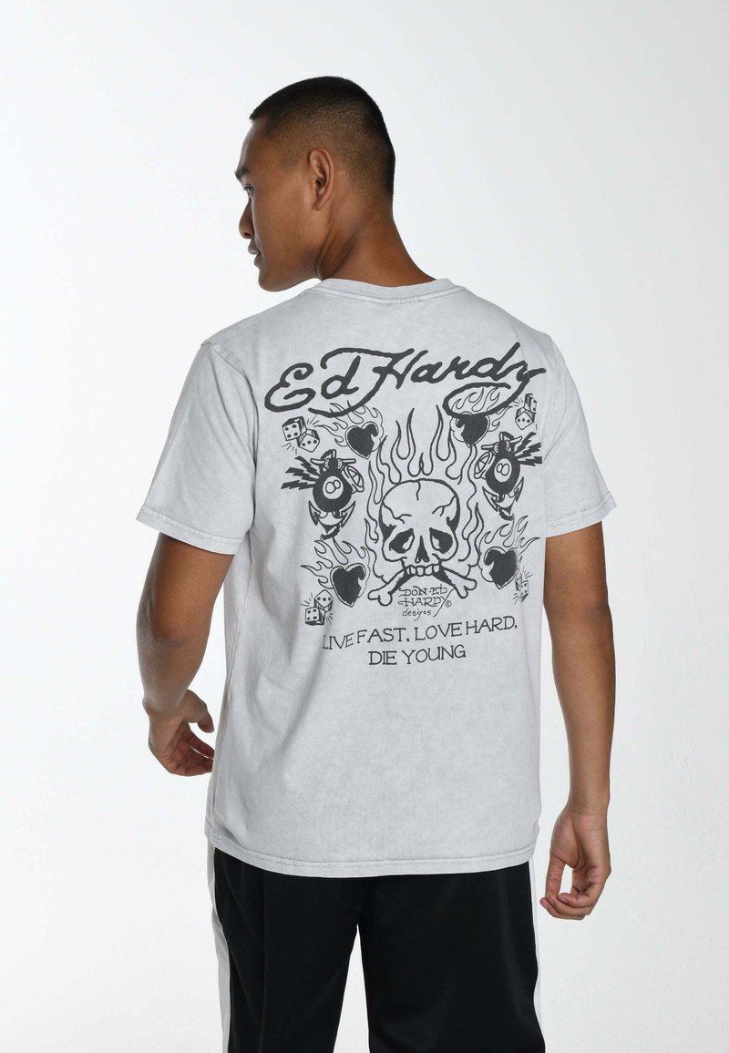 Camiseta gris con diseño gráfico negro en la parte posterior, que incluye una calavera, llamas y dados. Mangas cortas, cuello redondo y corte relajado.