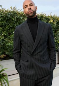 Traje gris oscuro de rayas de doble botonadura con un acabado texturizado, combinado con un ajustado cuello alto negro, de pie frente a un fondo de follaje verde.