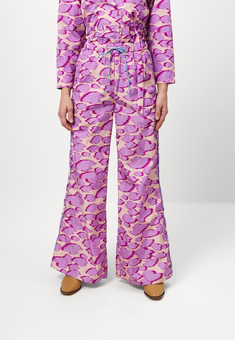 Personne portant un pantalon taille haute à jambes larges avec un motif abstrait rose et violet et un cordon bleu, associé à des chaussures beige.