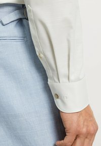 Weißes Langarmhemd mit glatter Textur und geknöpften Manschetten. Hellblaue Hose mit subtiler Textur und Seitentasche.