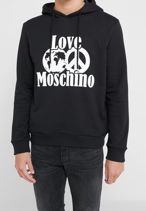 Persoon die een zwarte hoodie draagt met witte tekst "Love Moschino", een wereldbol en een vredessymbool op de voorkant, gecombineerd met donkere jeans.