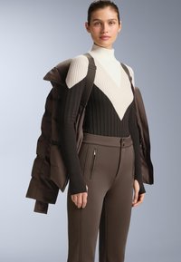 Čierny a krémový pletený sveter s rebrovanou textúrou, s V-vzorom. V kombinácii s hnedými úzkymi nohavicami a hnedou prešívanou bundou.