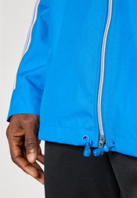 Blå jacka med dragkedja fram, med två blå snörstoppare vid fållen. Materialet verkar slätt och lätt med subtil textur.