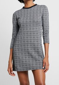 Robe à carreaux noir et blanc avec des manches longues, un col rond et une coupe ajustée. Texture lisse et accents de design minimalistes.