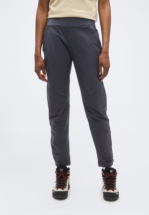 Personne portant un pantalon gris foncé avec une ceinture ajustée et des chaussures de randonnée beiges avec des lacets rouges, debout devant un fond uni.