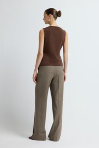 Donna con capelli raccolti a chignon, indossa un top senza maniche marrone, pantaloni a palazzo beige a quadri, e orecchini a cerchio dorati, in piedi su uno sfondo semplice.