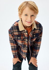 Flanellen shirt met een marineblauw, oranje en crèmekleurig ruitpatroon, beige ribstof kraag en twee voorkruiszakken. Model draagt een oranje shirt eronder.