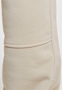 Beige jogger-housut, jotka on valmistettu pehmeästä, teksturoidusta kankaasta. Ominaisuuksiin kuuluu istuva muotoilu, jossa on näkyvä sauma ja hillitty resori nilkassa.
